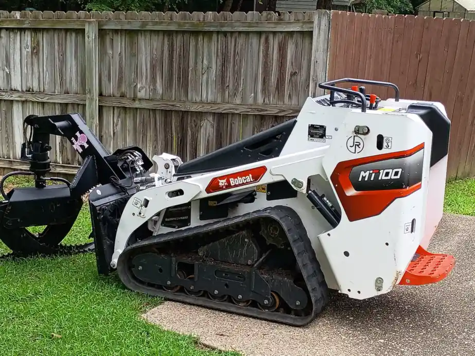 The white JR Tree Service Bobcat MT100 mini track loader with the JR circle decal on its side, grapple attached, on a lawn beside a wood fence