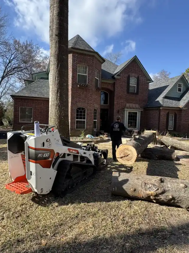 The mini track loader beside cut logs in front of a brick two-storey house