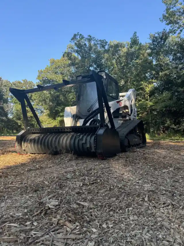 A forestry mulcher head on a track loader sitting on freshly mulched ground with pines behind